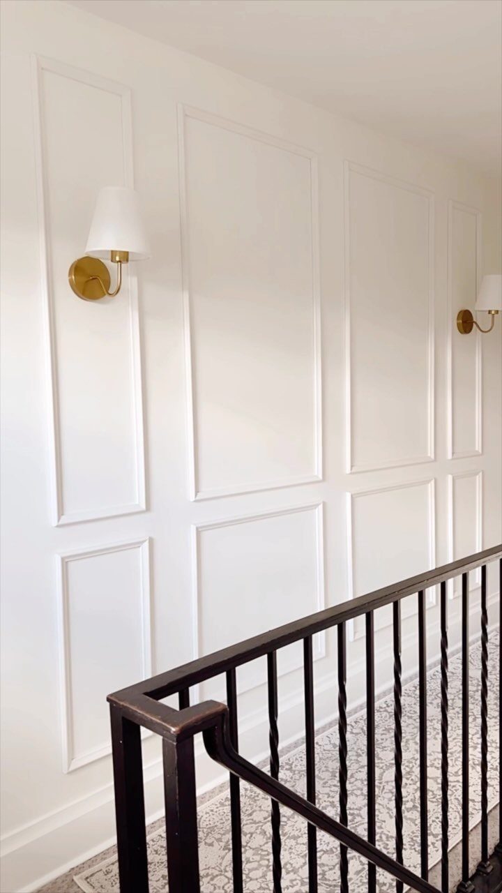 10 Interior Walls Ideas for 2026: Colors, Textures & Clever Details 8 White paneled walls featuring two gold metallic sconces with white lampshades near a staircase.