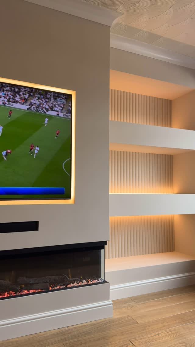 Recessed television with warm backlight above a fireplace and illuminated built-in shelving units.