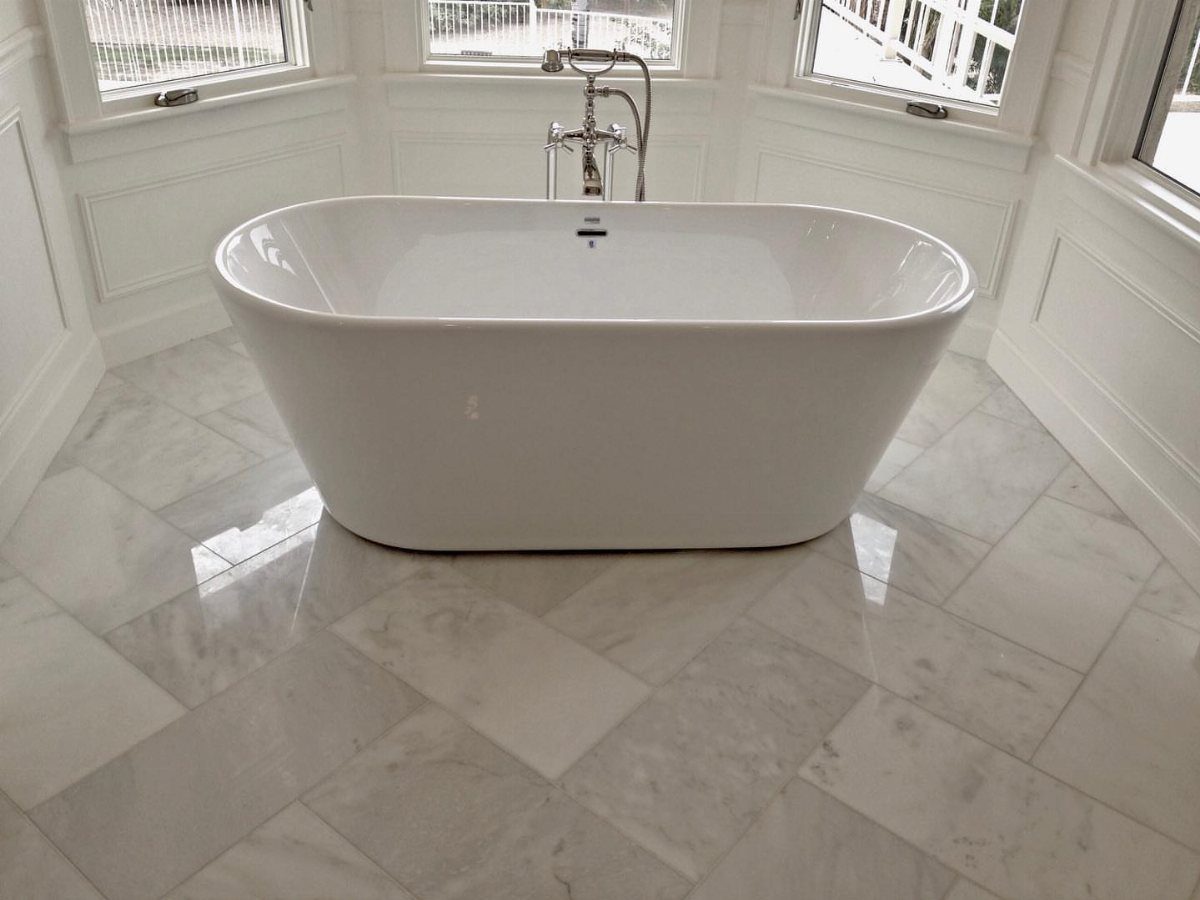 White bathtub centered on a light gray and white marble tile floor in a bright bathroom.