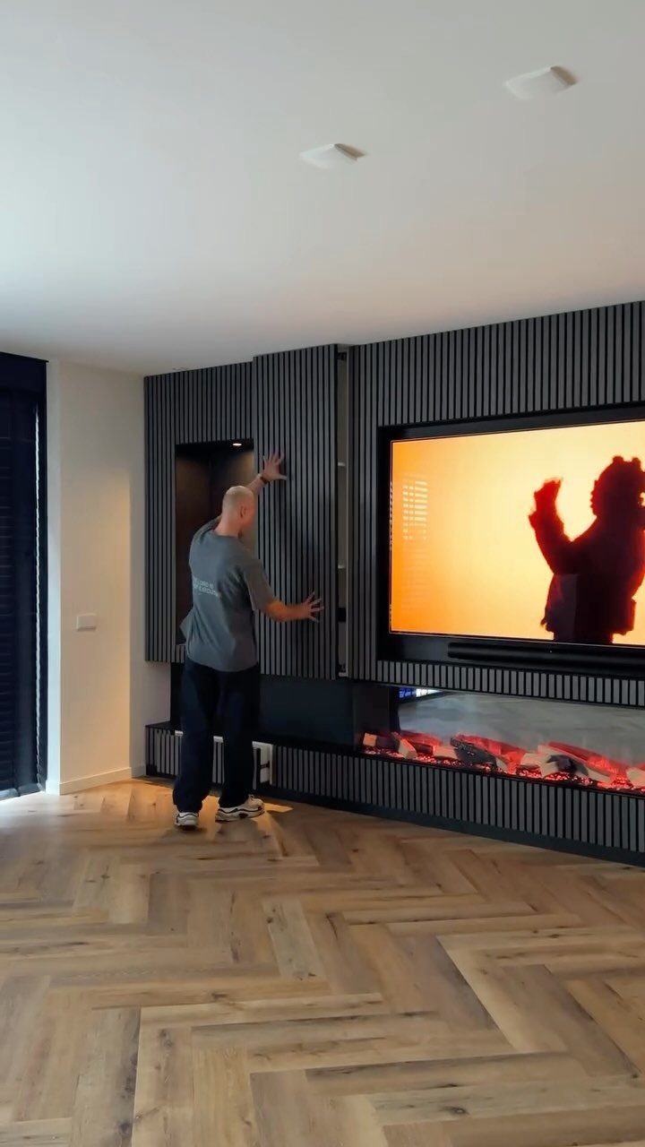A man stands beside a modern dark grey slatted media wall with a television and fireplace.
