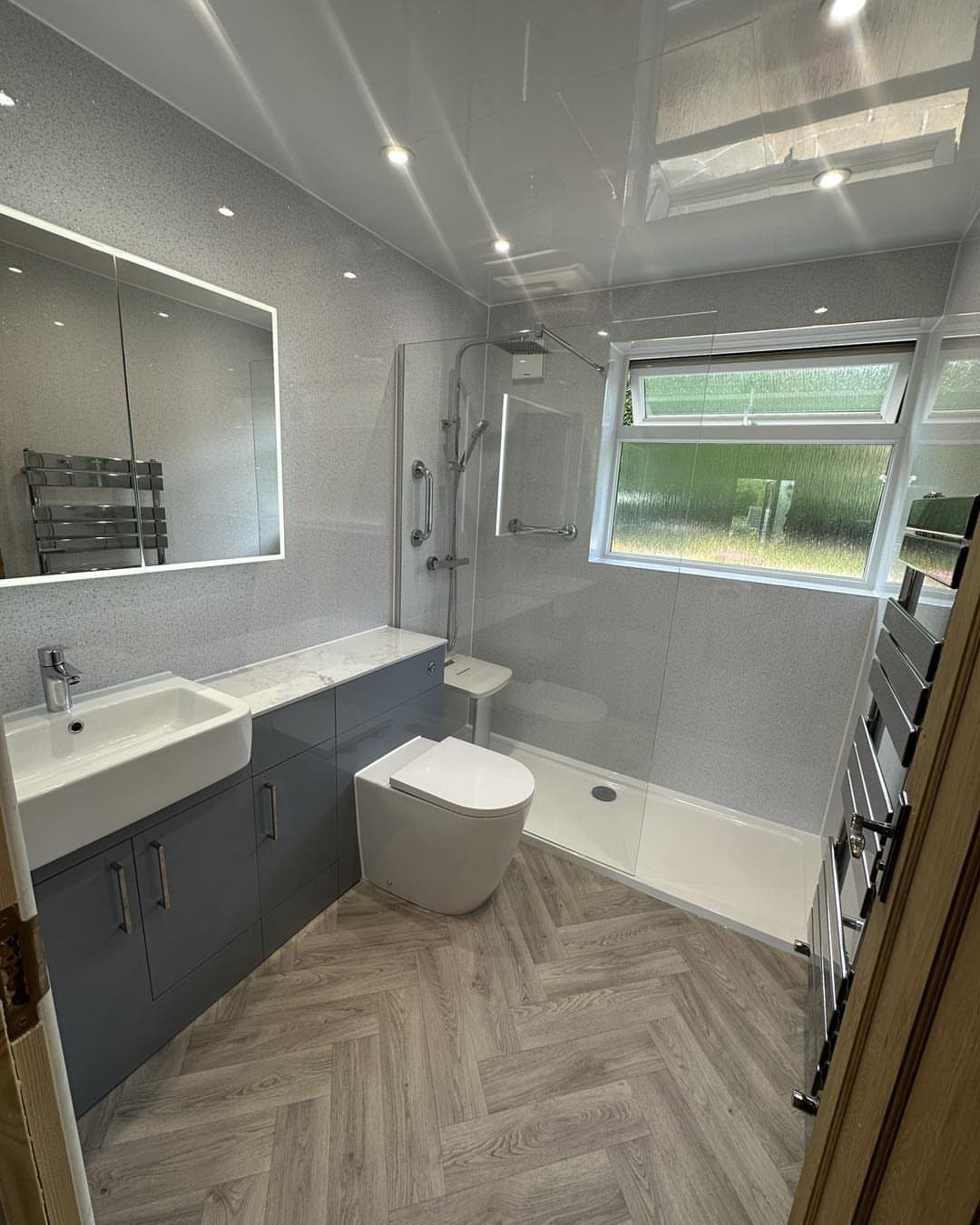 Modern bathroom with light grey glossy wall panels, blue vanity, and herringbone wood flooring.