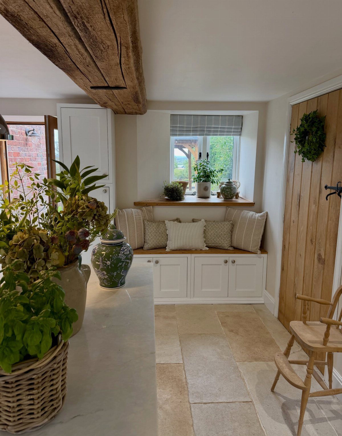 29 Rustic Farmhouse Kitchen Ideas That Feel Collected, Not Cataloged 28 White built-in window seat with cushions and potted plants under a wooden ceiling beam.