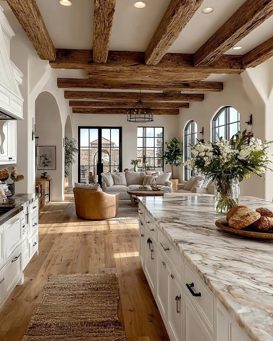 29 Rustic Farmhouse Kitchen Ideas That Feel Collected, Not Cataloged 25 White kitchen cabinets and marble countertops under rustic wooden ceiling beams in an open living space.