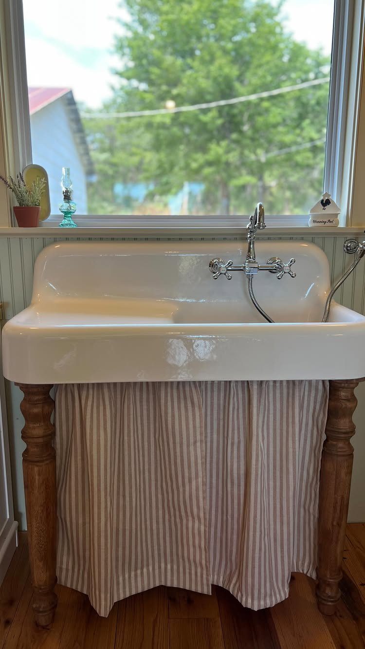 29 Rustic Farmhouse Kitchen Ideas That Feel Collected, Not Cataloged 1 A white ceramic farmhouse sink with a striped fabric skirt and wooden legs by a window.