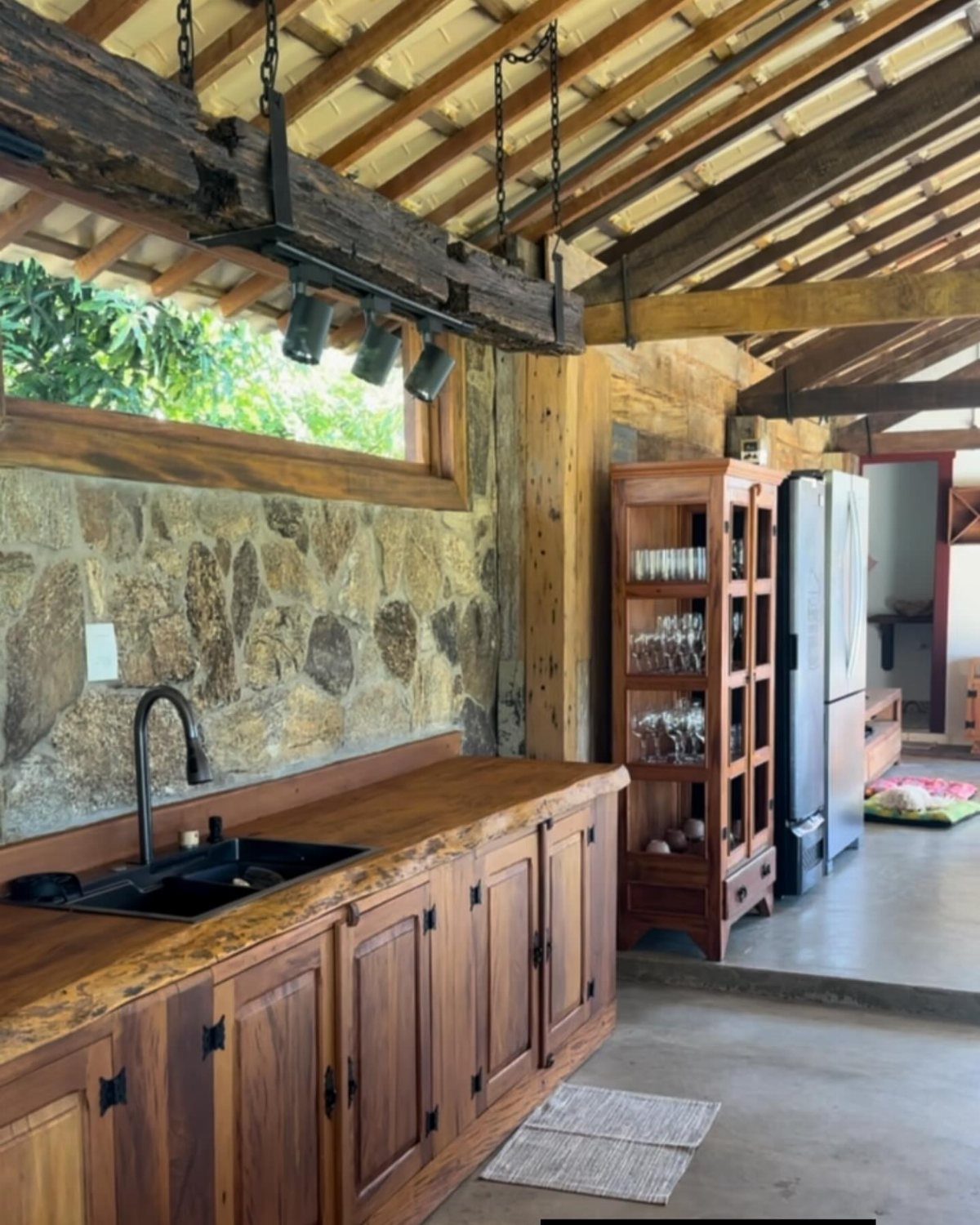 29 Rustic Farmhouse Kitchen Ideas That Feel Collected, Not Cataloged 14 Wooden kitchen cabinets with a dark sink and stone wall under a beamed ceiling.