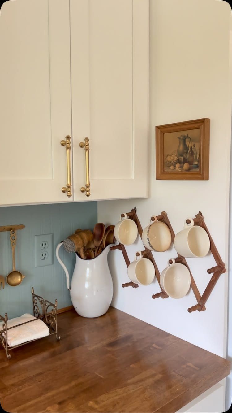 29 Rustic Farmhouse Kitchen Ideas That Feel Collected, Not Cataloged 16 Cream cabinets with gold vintage-style handles sit above a wooden countertop and mug rack.