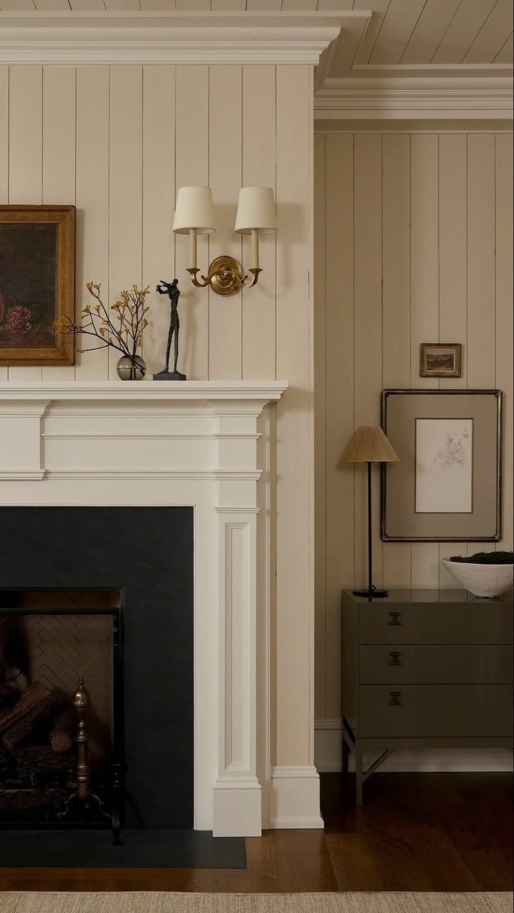 A white decorative fireplace mantel against a cream wood paneled wall with gold sconce lighting.