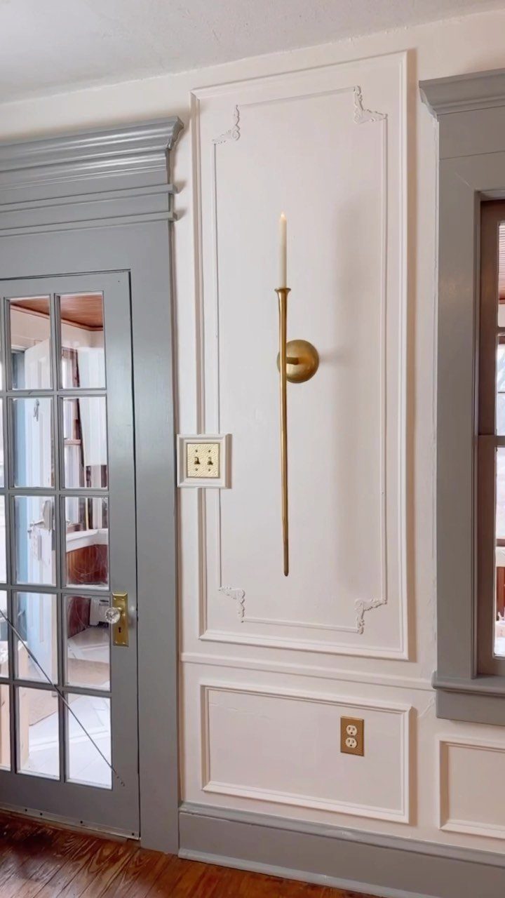 White decorative wall paneling with a gold sconce next to a grey door and window frame.