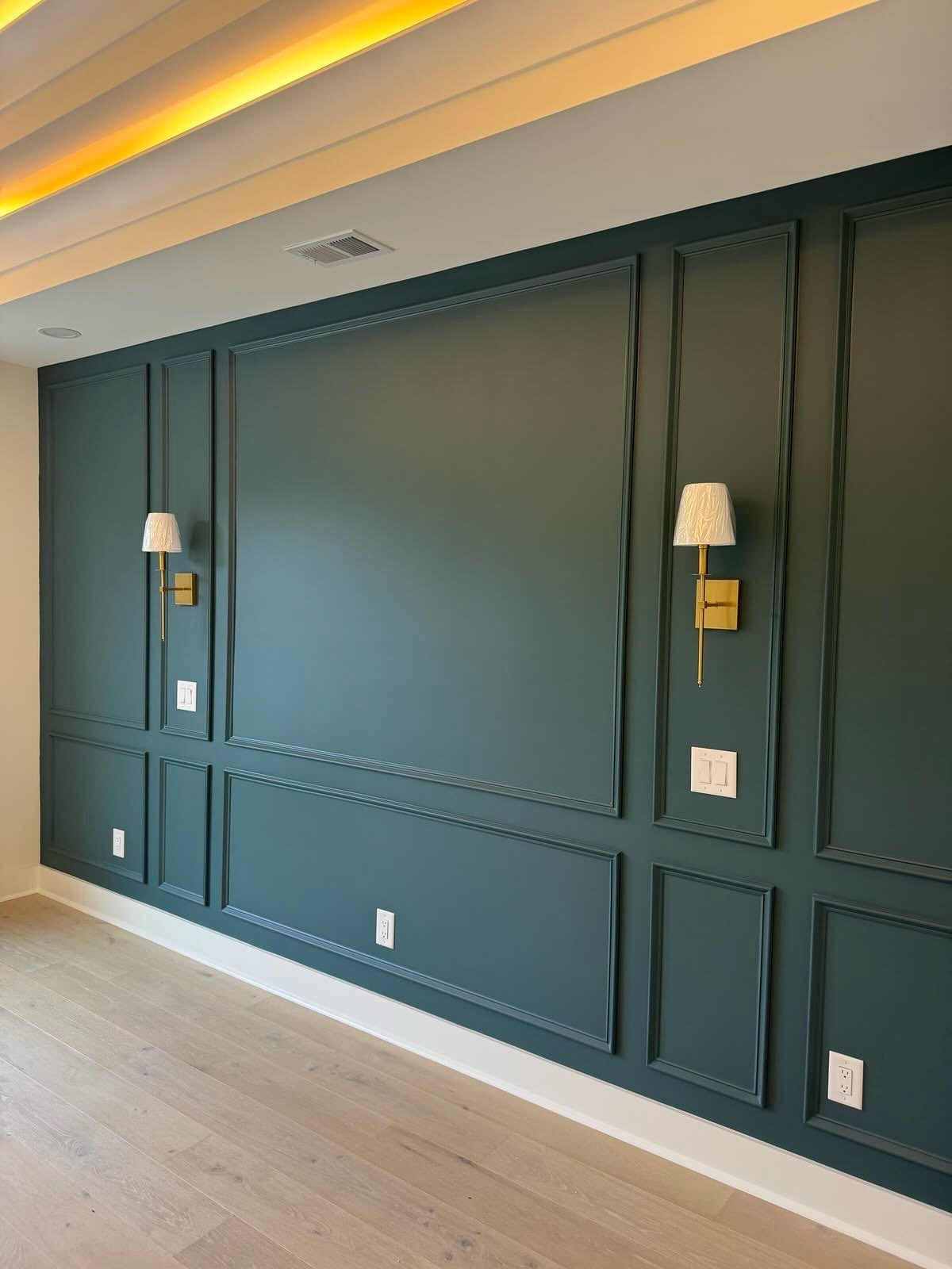 Dark teal paneled wall with decorative molding, gold sconces, and light wood flooring.