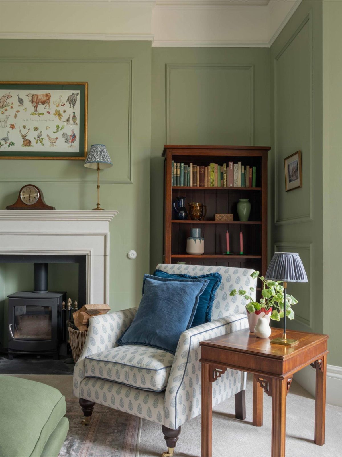 Sage green paneled walls in a living room with a white fireplace and patterned armchair.