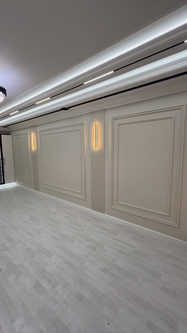 23 Wall Decor Design Ideas (Gallery Walls, Moss Walls, Rattan) 2 Two glowing vertical wall sconces mounted on a white paneled wall with decorative molding.