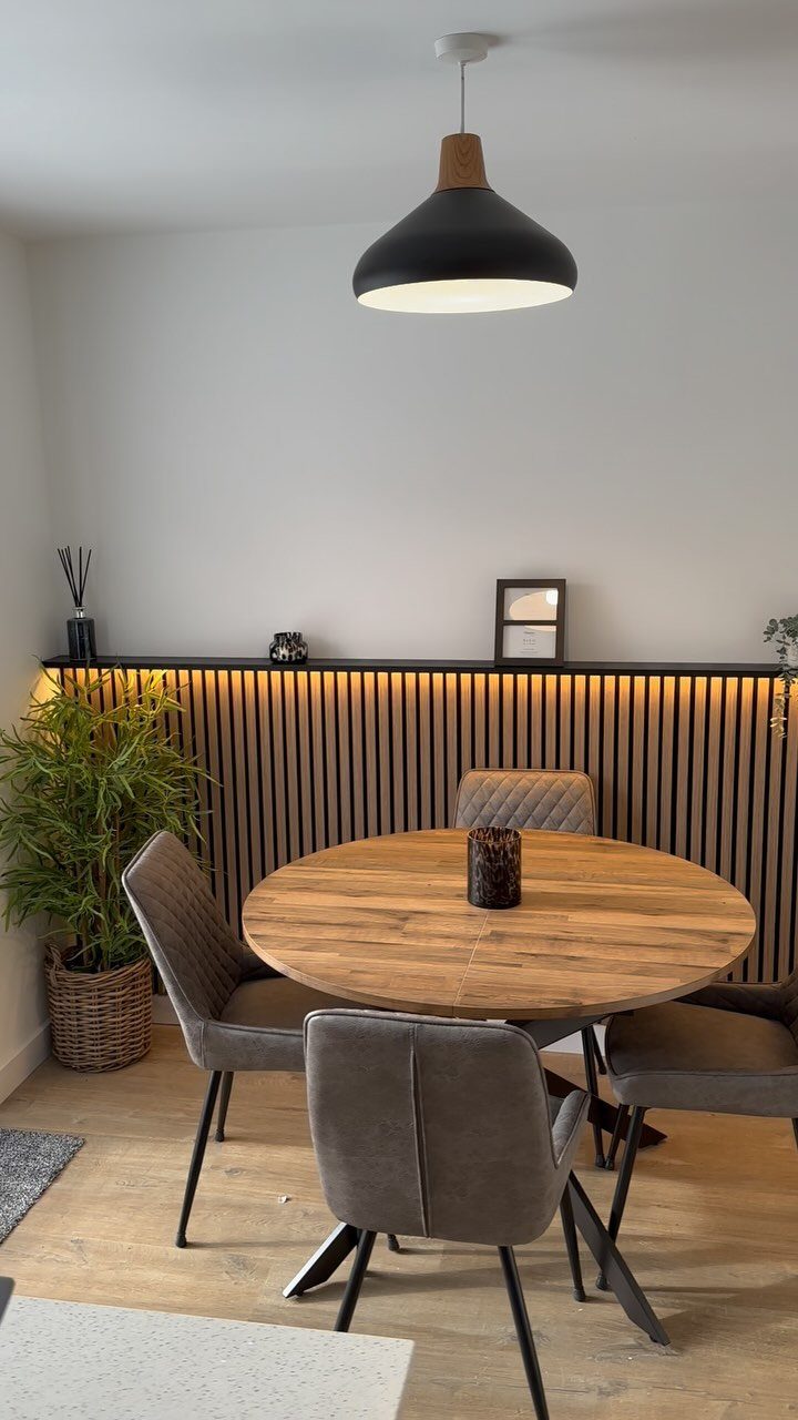 Wooden slat wall with warm LED lighting behind a round dining table and grey chairs.